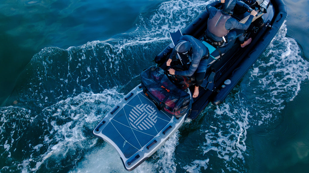 Aerial view of two spearfishers in wetsuits aboard a moving rigid inflatable boat featuring a Waihana-branded stern platform and the Expedition Load Out gear bag, surrounded by churning ocean wake.